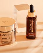 Lancôme Absolu Cream and L’Oréal Paris Facial Tanning Drops photographed on a peach background.