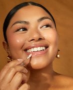 Person smiling with a natural makeup look and hair pulled back off of their face applies clear lip gloss to their lips